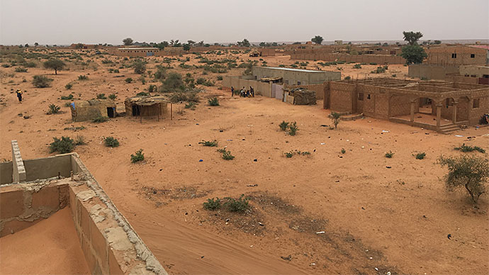 Afrikansk steppelandskap med noen trær, busker og mye sand. Bildet viser noen enkle huskonstruksjoner noen mennesker langt unna.