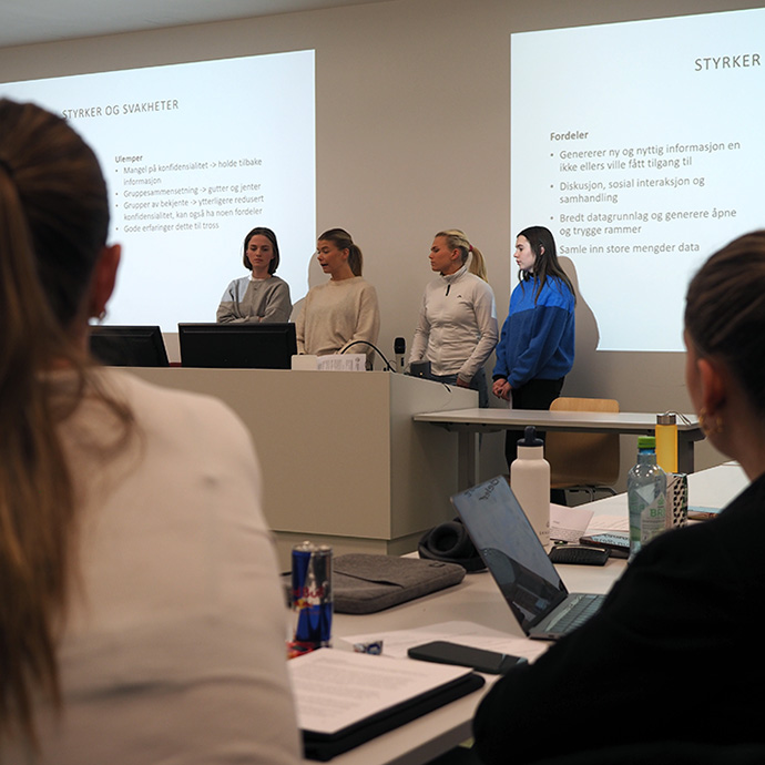 Studentene står foran sine medstudenter og presenterer sitt prosjekt som vises på PowerPoint-presentasjon i bakgrunnen.