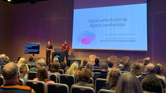 Ingunn Ikdahl står bak en talerstol og holder en presentasjon. På lerrett bak henne vises et lysbildeark med teksten "Digital velfersstat og digitalt utenforskap". Ved siden av henne på scenen står en tegnspråktolk, samt en skjerm brukt til skrivetolkning.